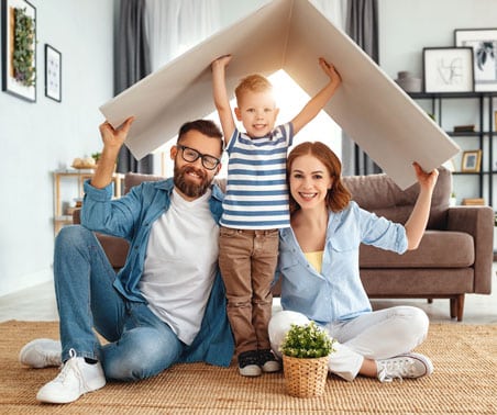 A smiling man, woman, and young boy sit on a living room floor, holding a cardboard roof shape above them, symbolizing family unity.
