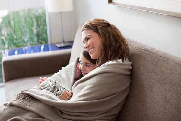 A woman and young girl sit together on a couch, smiling under a beige blanket with sunlight streaming through the window behind them.