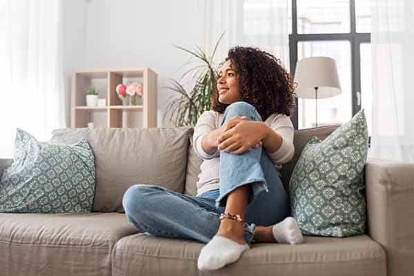 A woman sits on a sofa, smiling and gazing out the window, dressed in a white top and jeans. The bright room has plants and cushions.