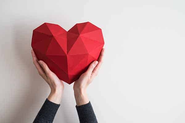 A person holding a large geometric red heart with both hands against a plain white background.