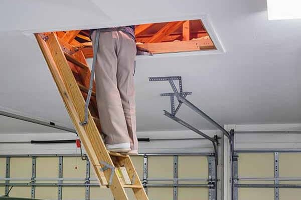 A person stands on a wooden attic ladder in a garage, upper body in the attic and legs visible. Garage door and ceiling behind.