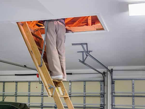 A person stands on a wooden attic ladder in a garage, upper body in the attic and legs visible. Garage door and ceiling behind.