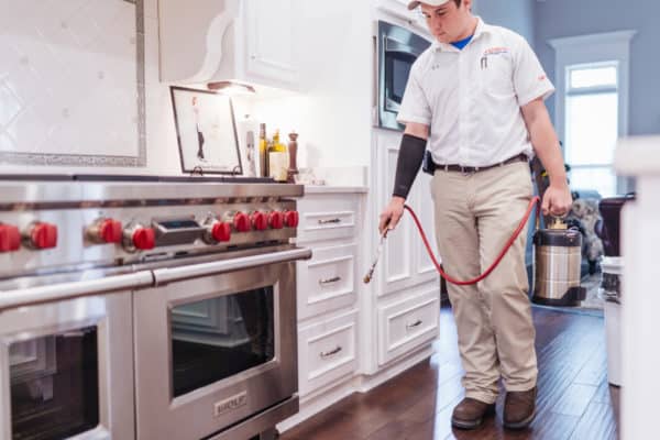 A pest control technician in uniform sprays baseboards of a modern kitchen with a stainless steel stove, using a metal canister.