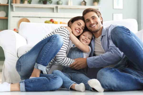 A smiling family of three sits together on the floor by a white couch, hugging and posing happily in a bright, cozy living room.