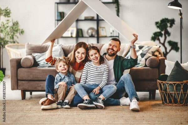 A happy family of four sits on their living room floor, smiling and holding a cardboard roof above their heads in front of a couch.