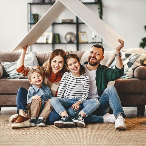 A happy family of four sits on their living room floor, smiling and holding a cardboard roof above their heads in front of a couch.