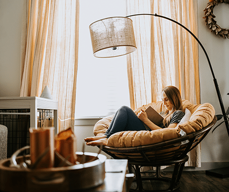 A woman reads in a round cushioned chair by a large window with sheer curtains. Warm light fills the cozy room with simple decor.