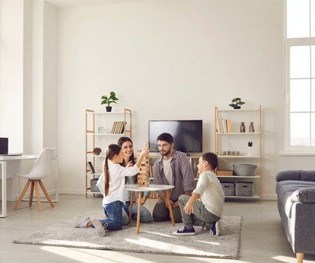 A family of four sits on the floor in a bright living room, playing Jenga on a small table with shelves, a TV, and large windows behind.