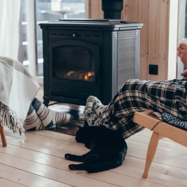 Two people in cozy pajamas sit by a wood stove, one with a mug. A black cat lounges nearby on the floor. The scene feels inviting.