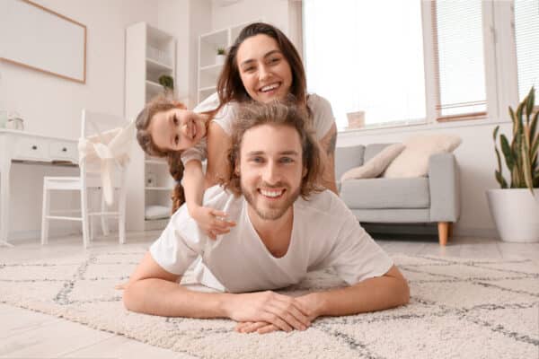 A happy family in white shirts lies playfully together on the bright living room floor, with sofa, desk, and plants in the background.