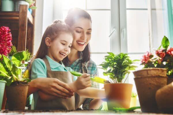 A smiling woman and young girl in aprons tend to potted plants indoors, sunlight streaming in, with a table of greenery and tools.