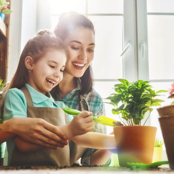 A smiling woman and young girl in aprons tend to potted plants indoors, sunlight streaming in, with a table of greenery and tools.
