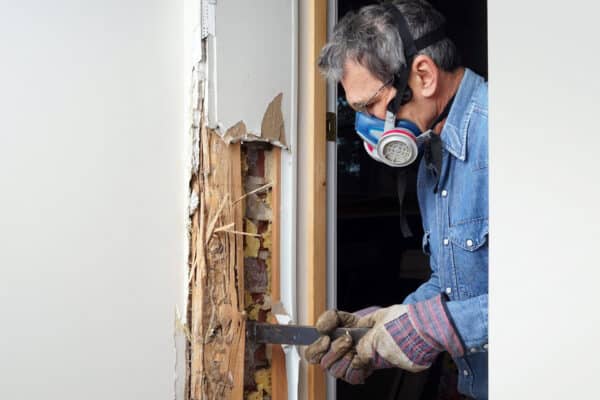 A person in gloves, denim shirt, protective eyewear, and face mask uses a tool to remove damaged wood from a door frame, exposing insulation.