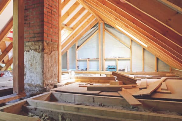 Unfinished attic with exposed beams and brick chimney, construction materials and planks scattered, light entering from far windows.