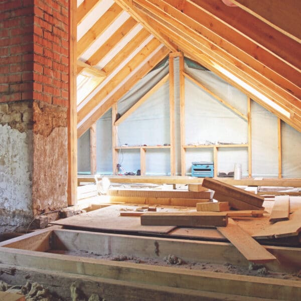 Unfinished attic with exposed beams and brick chimney, construction materials and planks scattered, light entering from far windows.