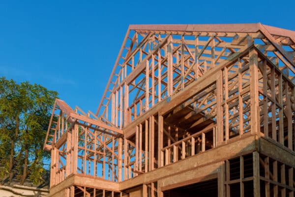 A partially built wooden house frame stands under a clear blue sky, with trees in the background and exposed beams and trusses visible.