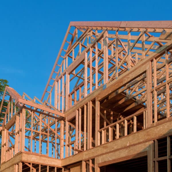 A partially built wooden house frame stands under a clear blue sky, with trees in the background and exposed beams and trusses visible.