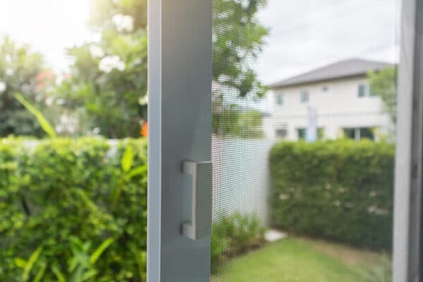 Close-up of a sliding window with mesh screen, partly open, showing green bushes and a blurred house outside on a sunny day.