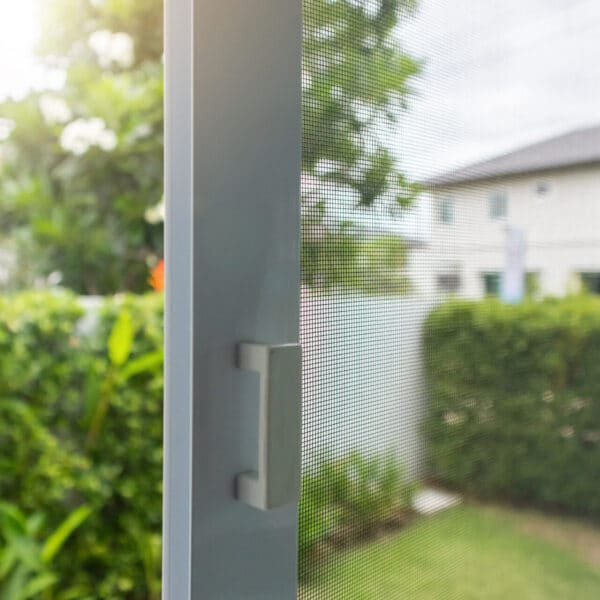 Close-up of a sliding window with mesh screen, partly open, showing green bushes and a blurred house outside on a sunny day.