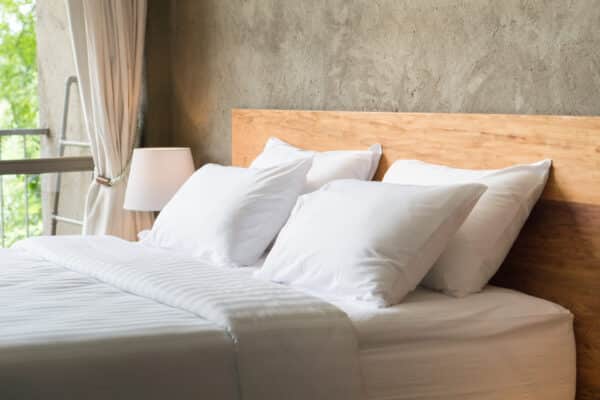 A neatly made bed with white linens and four pillows rests by a wooden headboard, lamp, and bright window with airy curtains.