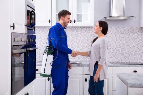 A pest control technician in blue overalls shakes hands with a woman in a modern kitchen, holding a pest spray tank.