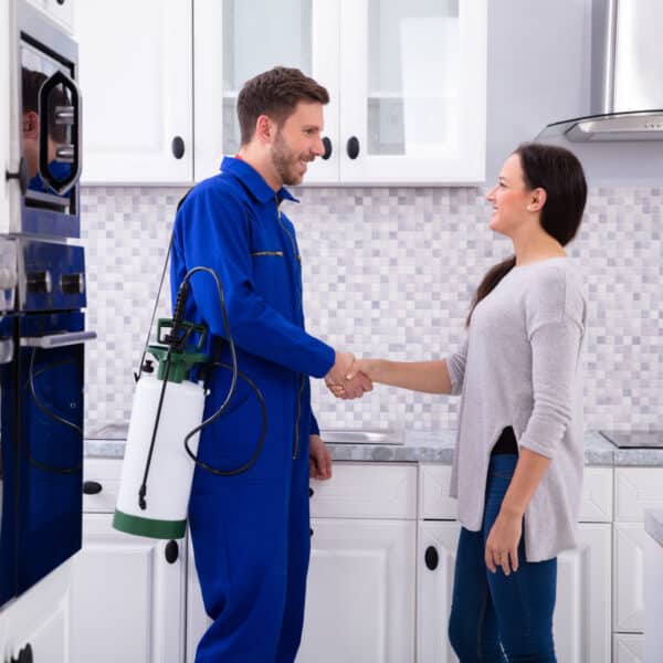 A pest control technician in blue overalls shakes hands with a woman in a modern kitchen, holding a pest spray tank.