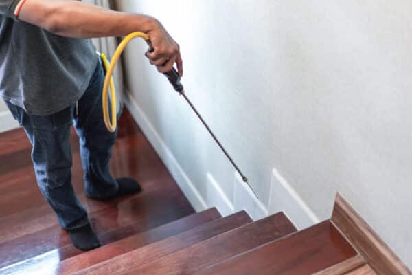 A person sprays insecticide or pesticide along the base of a wall next to wooden stairs, using a long nozzle and yellow hose.