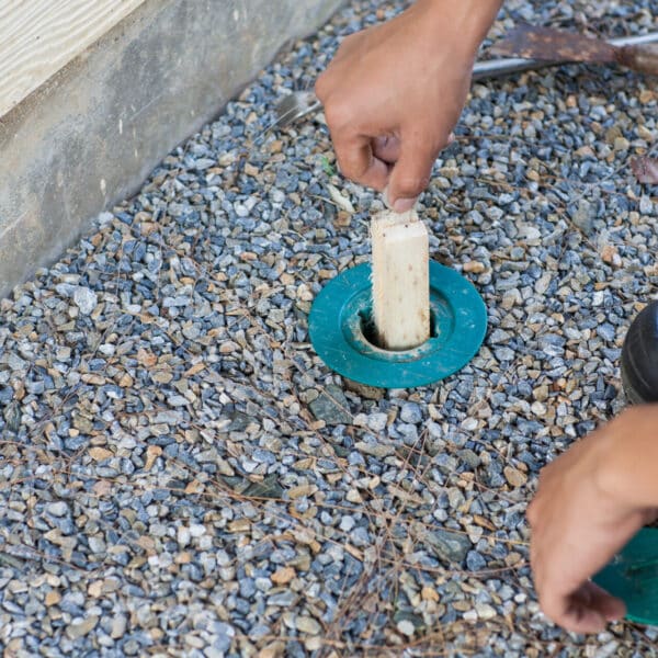 A person inserts a wooden stick into a green plastic ground cover on gravel near a building’s foundation, with tools and a shoe nearby.
