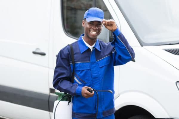 Smiling pest control worker in blue uniform and cap stands by a white van, holding equipment and touching the brim of his cap.