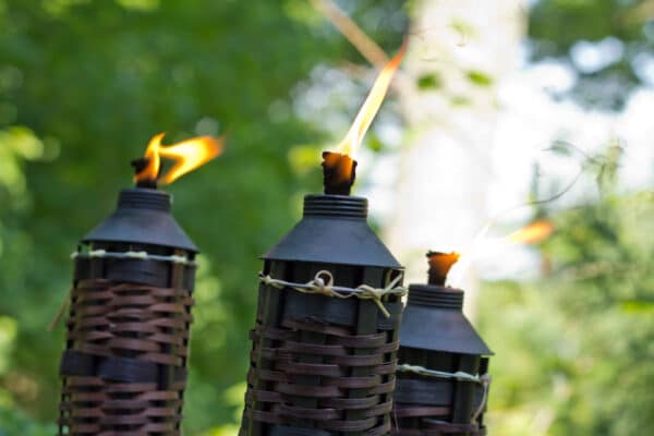 Three tiki torches with black metal tops and woven bamboo bodies burn with small flames, against a blurred green outdoor background.
