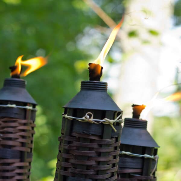 Three tiki torches with black metal tops and woven bamboo bodies burn with small flames, against a blurred green outdoor background.