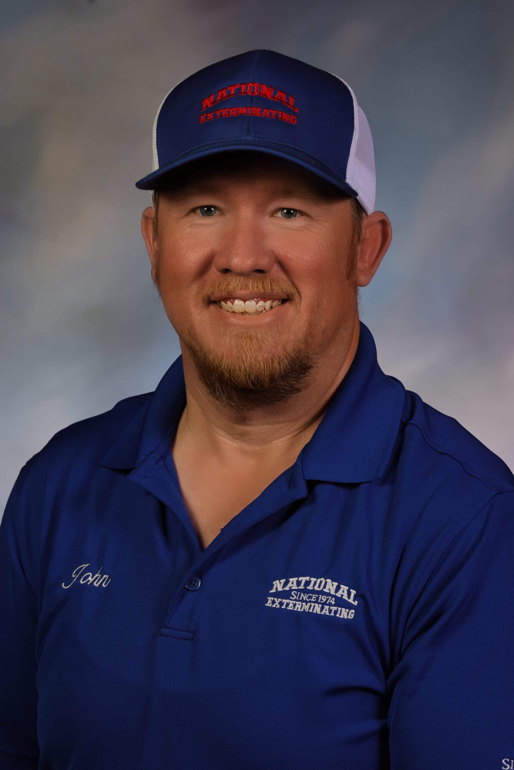 A man with a reddish beard wears a blue collared shirt embroidered with John and National Exterminating, and a matching cap. He is smiling against a soft, blurred background.