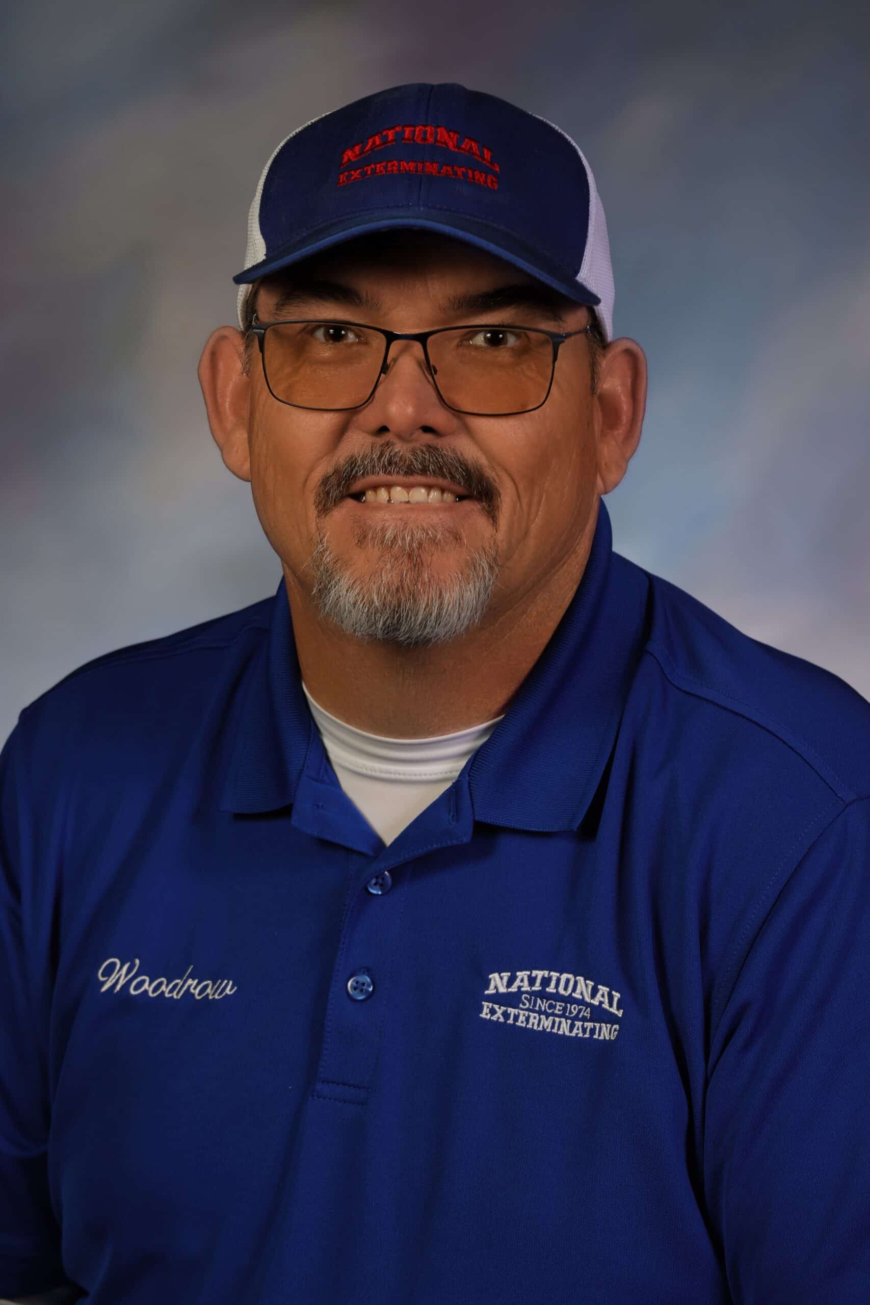 A man with glasses, a goatee, and a cap embroidered with National Exterminating smiles at the camera. He wears a blue collared shirt with National Exterminating and Woodrow embroidered on it.