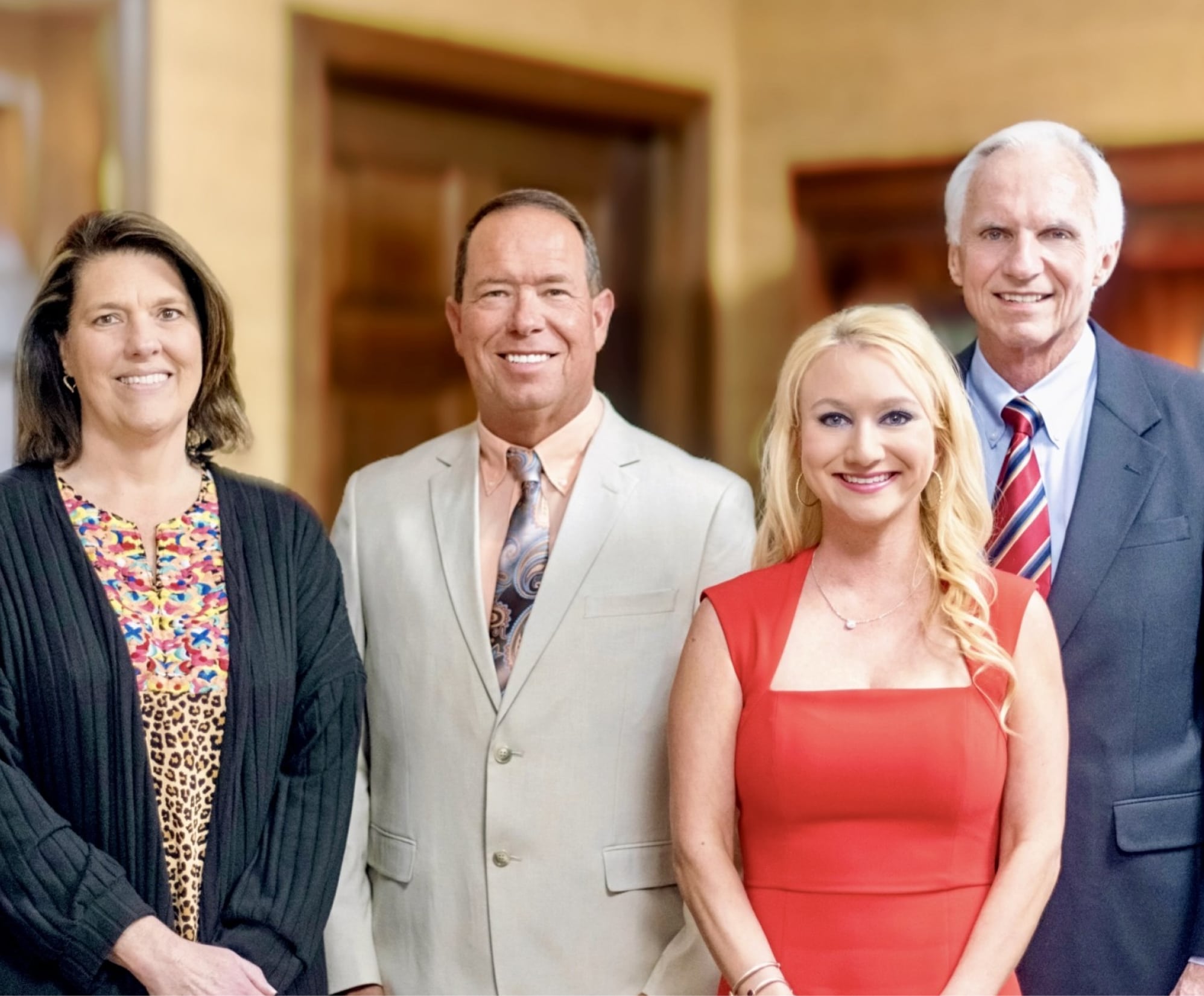 Four adults stand indoors, smiling at the camera. Two men in suits are between two women, one in black and the other in a red dress.