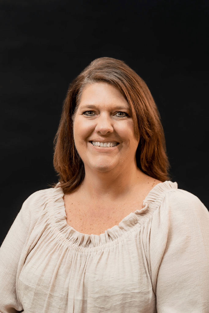 A woman with shoulder-length brown hair smiles at the camera, wearing a light ruffled blouse against a plain black background.