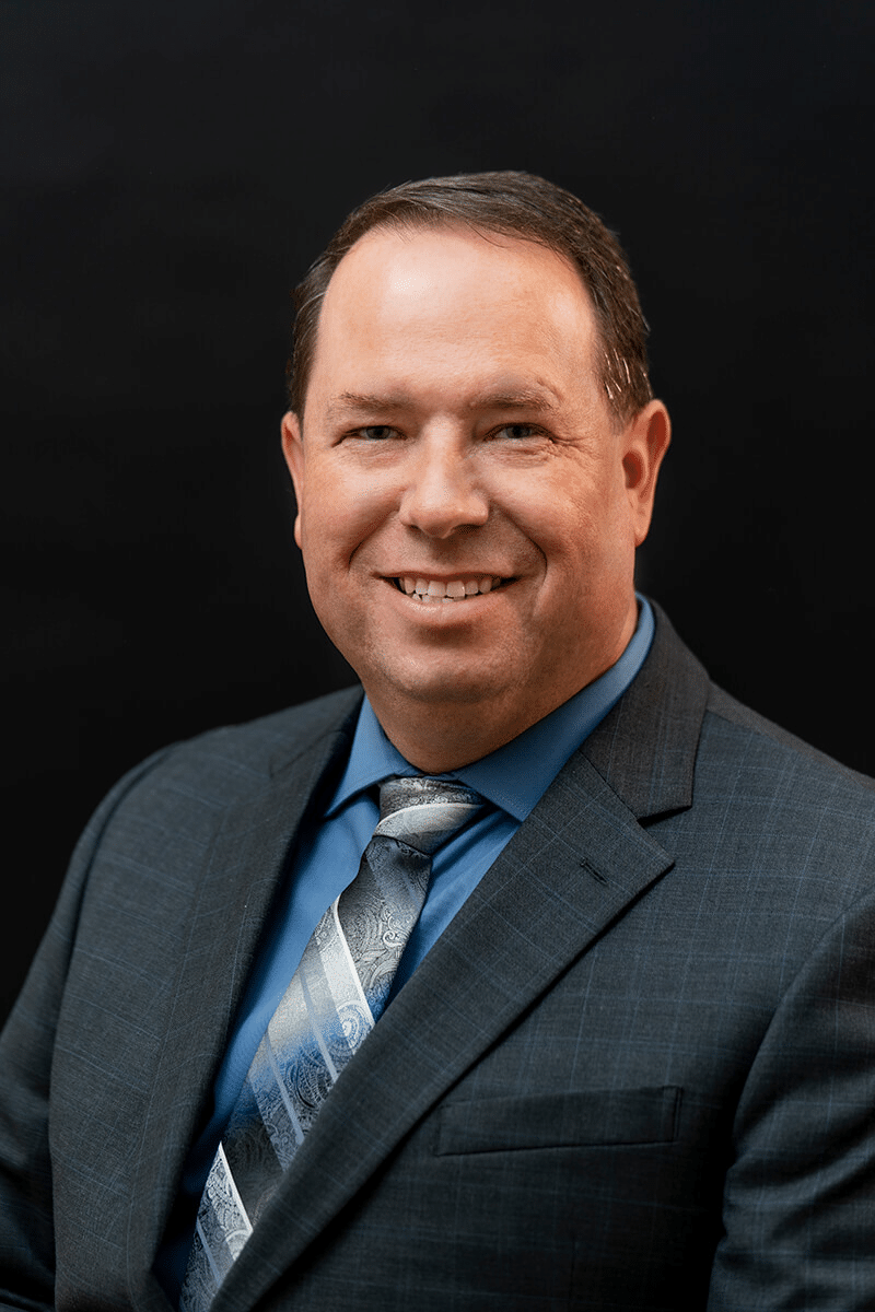 A middle-aged man in a gray suit, blue dress shirt, and patterned tie smiles at the camera against a plain black background.
