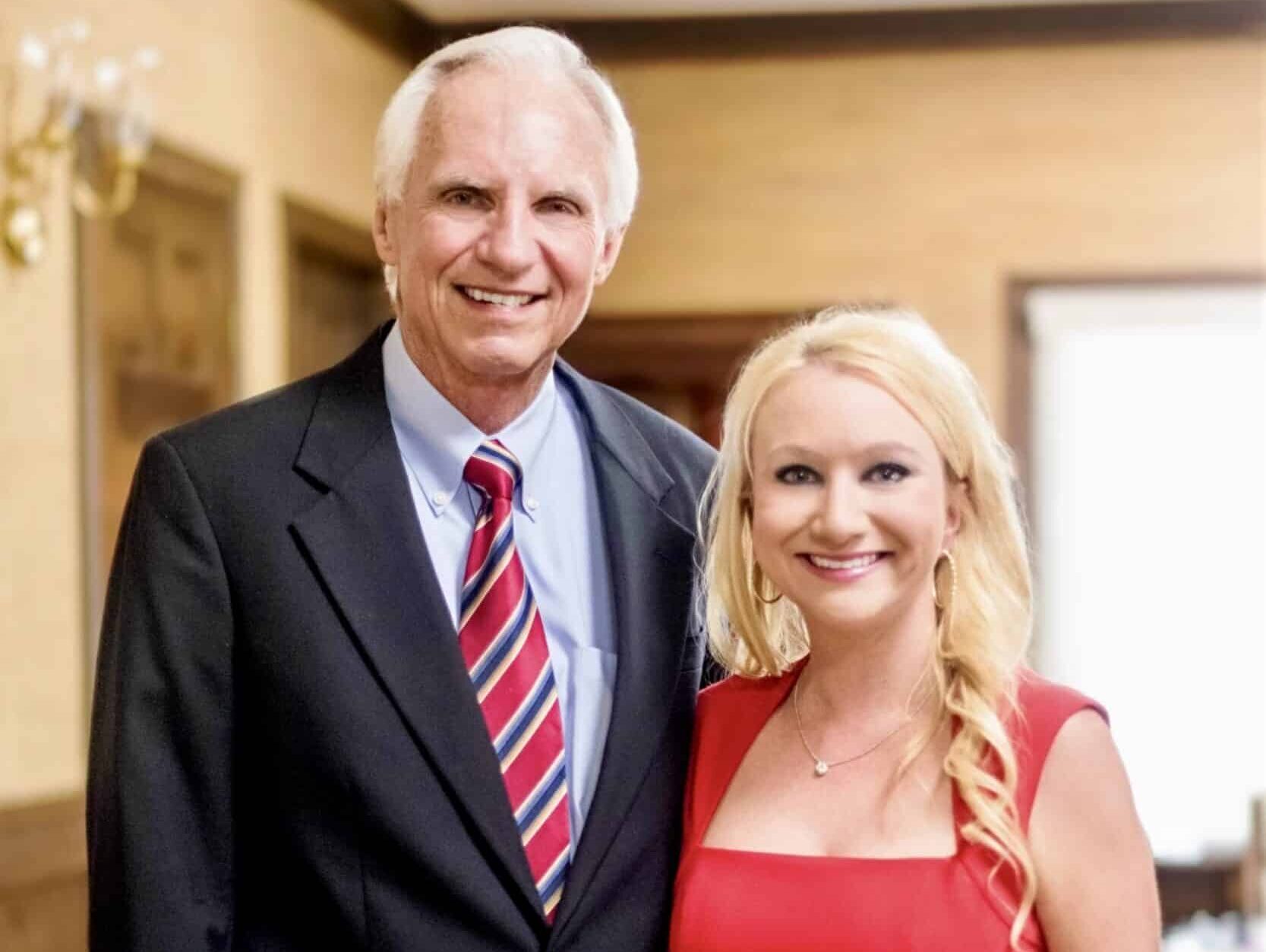 A smiling man in a dark suit and striped tie stands beside a smiling woman in a red dress in a room with wood paneling and pale walls.