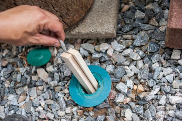 A hand opens a green plastic cap on a termite bait station set in gravel, exposing wooden monitoring stakes inside.
