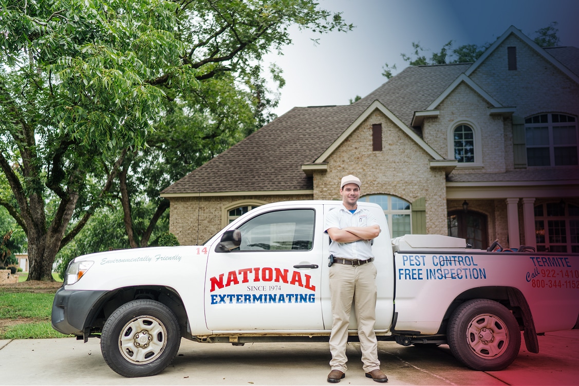 A pest control technician stands smiling with arms crossed in front of a National Exterminating truck by a suburban brick house with trees.