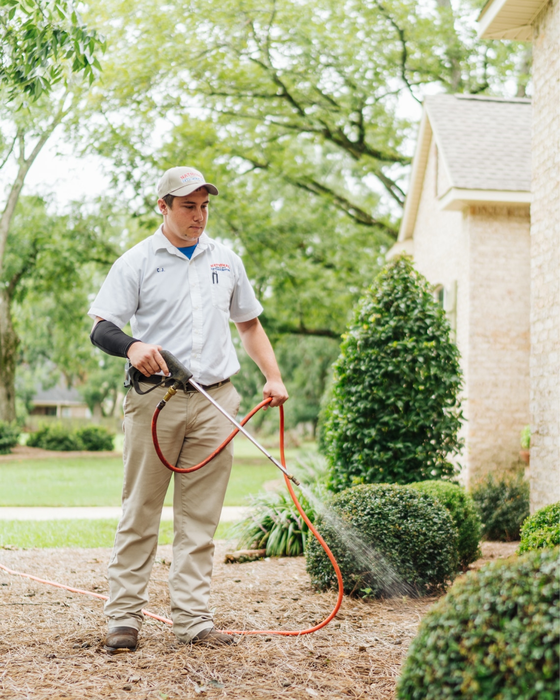 A pest control worker in uniform and cap sprays pesticide on shrubs by a house with a hose. Green bushes and trees are in the background.