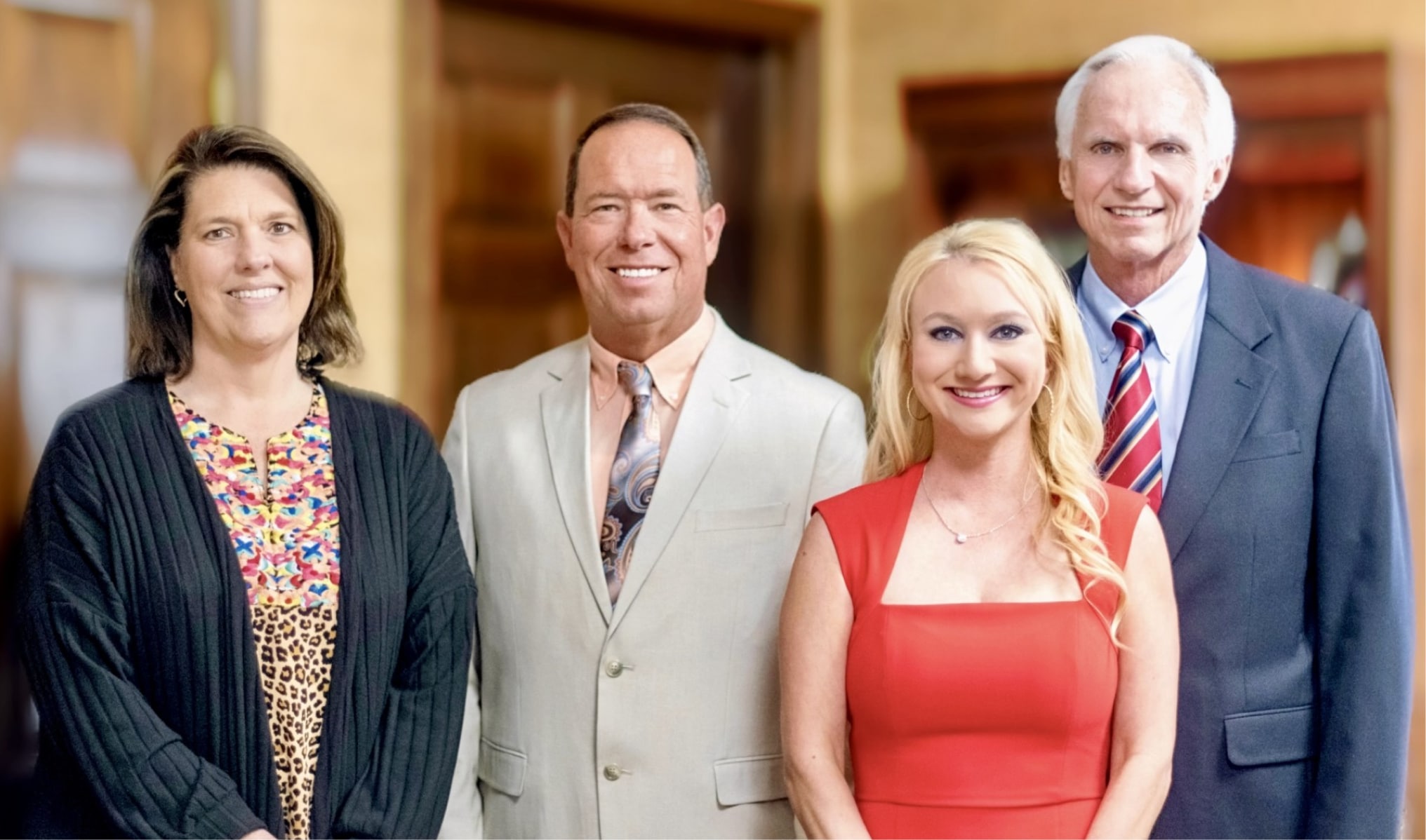 Four adults stand indoors, smiling at the camera. Two men wear suits; two women wear patterned tops, one in a red dress.
