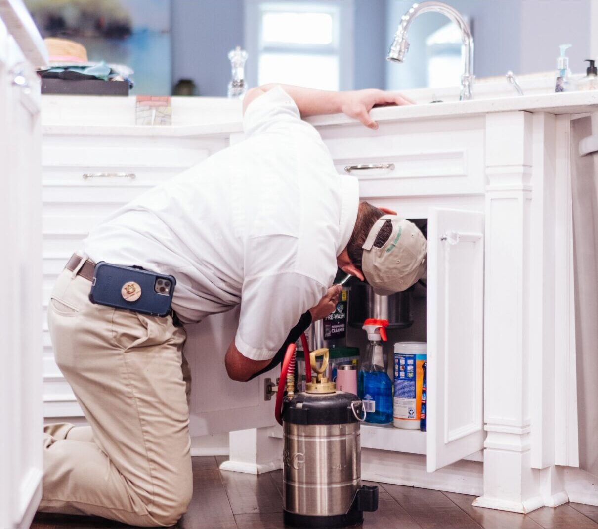A pest control technician in uniform kneels, checking under a kitchen sink with cleaning supplies, holding equipment, and wearing a cap.