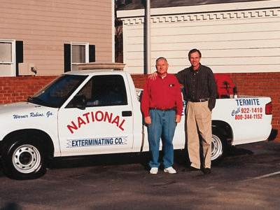 Two men stand smiling next to a white pickup truck labeled National Exterminating Co. and TERMITE, parked in front of a house. The truck has business phone numbers and location information written on it.