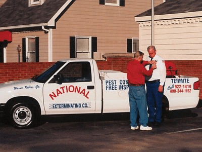 Two men stand next to a white pickup truck labeled National Exterminating Co. One pins something on the other's shirt. Pest control services are advertised.