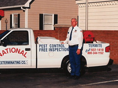 A man in a white uniform stands by a pest control truck with signage and phone numbers, parked near a brick wall and beige building.