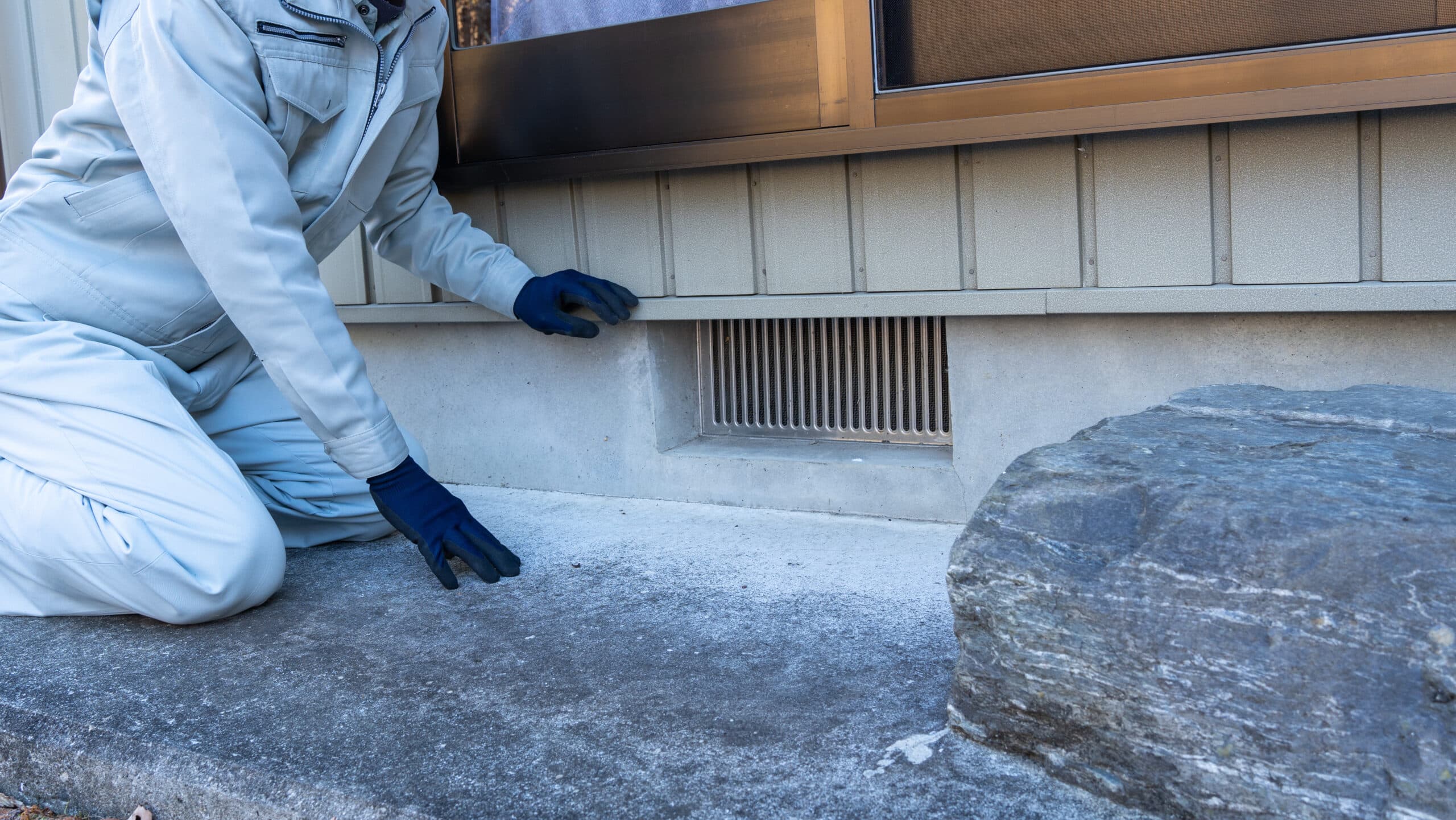 A person in light work clothes and dark gloves kneels by a building, inspecting a vent and white powder near the base. A rock is nearby.