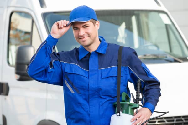 A smiling pest control worker in blue uniform and cap stands by a white van, holding a pesticide sprayer and touching his cap's brim.