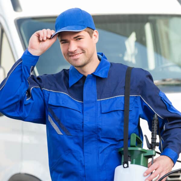 A smiling pest control worker in blue uniform and cap stands by a white van, holding a pesticide sprayer and touching his cap's brim.