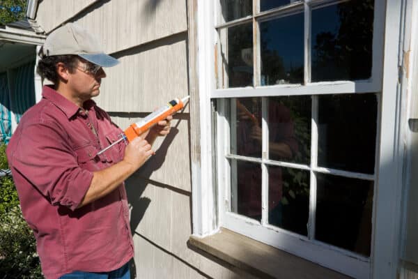 A person in a cap and sunglasses uses a caulking gun to seal an exterior window frame of a house on a sunny day.