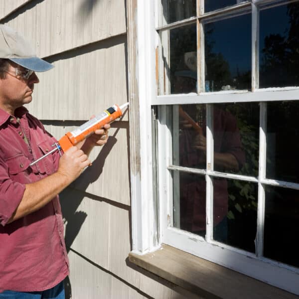 A person in a cap and sunglasses uses a caulking gun to seal an exterior window frame of a house on a sunny day.